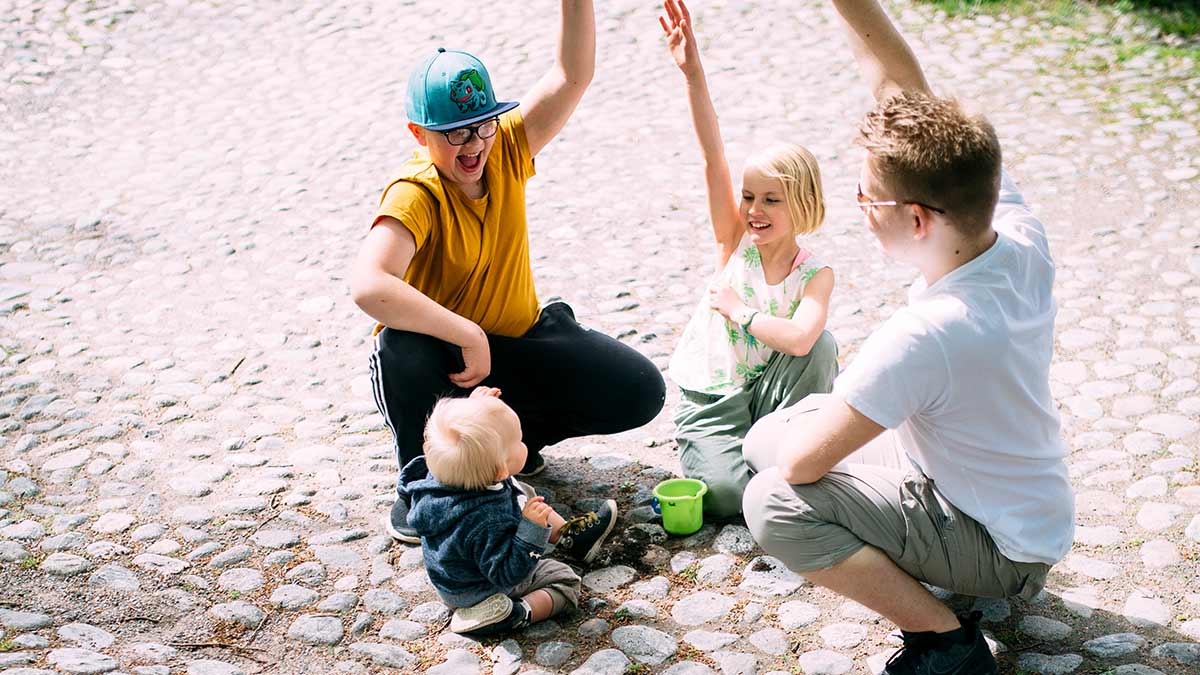 Taapero, nuori poika, nuori tyttö ja teini-ikäinen poika yhdessä nostamassa kädet ilmaan.