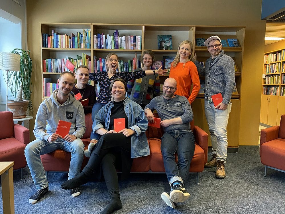 Nuoren seurakunnan veisukirjan toimittajat istuvat sohvalla. Osa heistä seisoo sohvan ympärillä. Kaikki pitelevät veisukirjoja käsissään.