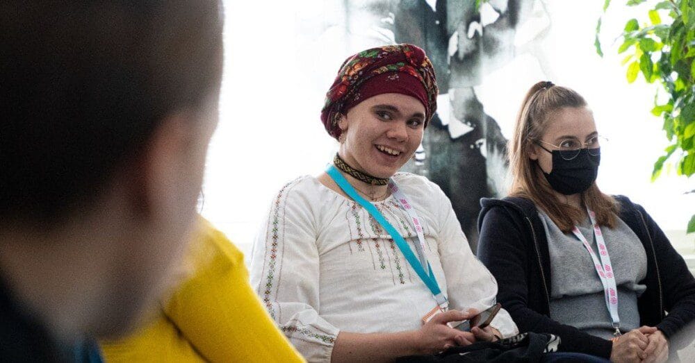 Kaksi nuorta istuu ikkunan edessä. Toinen keskustelee etualalla näkyvän henkilön kanssa ja hymyilee. Hänellä on puhelin kädessä. Vieressä istuva nuori katsoo eteenpäin keskittynyt ilme kasvoillaan,