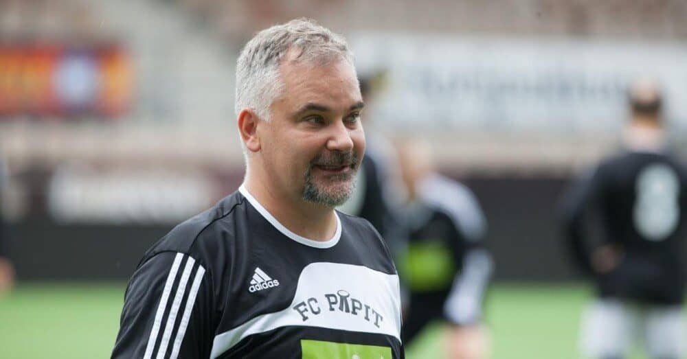 Miehellä on jalkapallopaita päällä ja taustalla näkyy sumeana muita jalkapallonpelaajia. Paidassa lukee FC papit. Katse on keskittynyt ja pieni hymy kasvoilla.