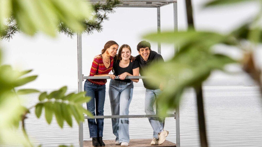 Kolme nuorta seisoo rannalla vierekkäin ja hymyilee. Tunnelma on kesäinen.