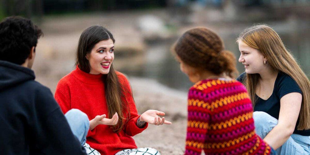 Neljä nuorta istuu kalliolla. Yksi heistä selittää voimakkaasti käsillään hymyillen muille jotakin.