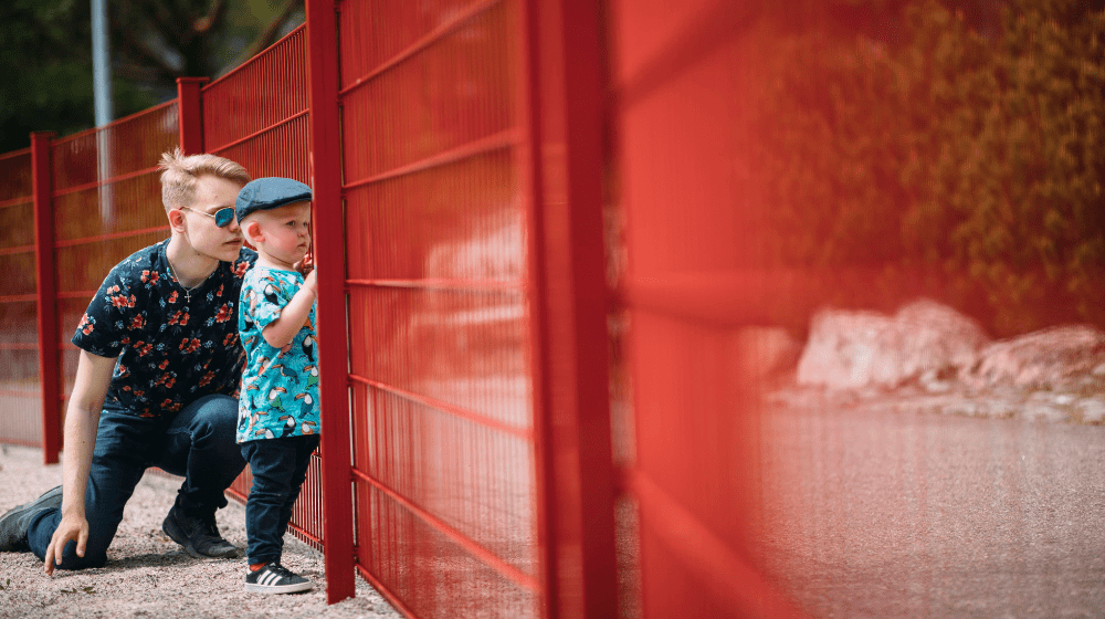 Mies ja pieni poika ovat aidan vieressä ja vaikuttavat tarkkailevan aidan raoista.