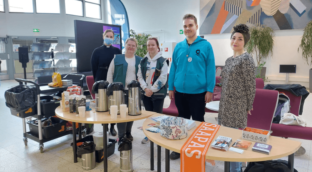 Viisi aikuista seisoo pöytien takana aulamaisessa tilassa. Pöydillä on esitemateriaaleja ja kahvitarvikkeita.