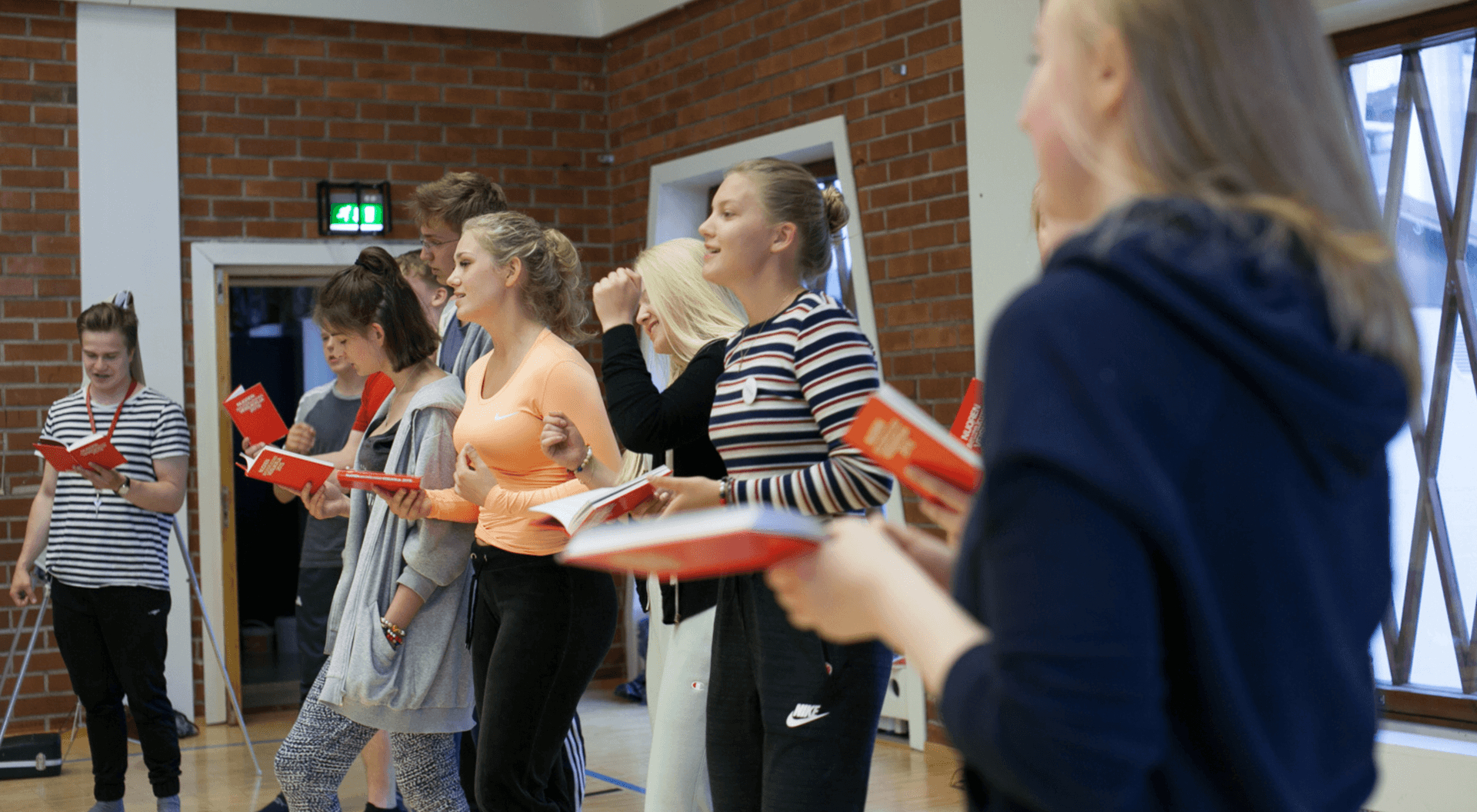 Nuoret seisovat ja laulavat virsikirjat käsissään. He ovat jumppasalin tyylisessä tilassa.