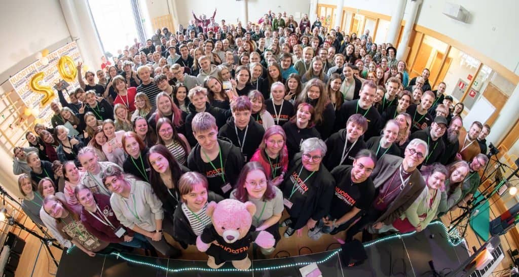 Suuri joukko nuoria ja aikuisia kokoontuneena yhteen liikuntasaliin. Kuva on otettu ylhäältäpäin, niin että ihmisjoukko näyttää pieneltä massalta ja kaikki katsovat ylöspäin kameraan. Yllään heillä on tavalliset, rennot vaatteet. Etualalla kaksi nuorta naisoletettua pitää suurta teddykarhua, jolla on yllään tulevaisuusseminaarin paita.