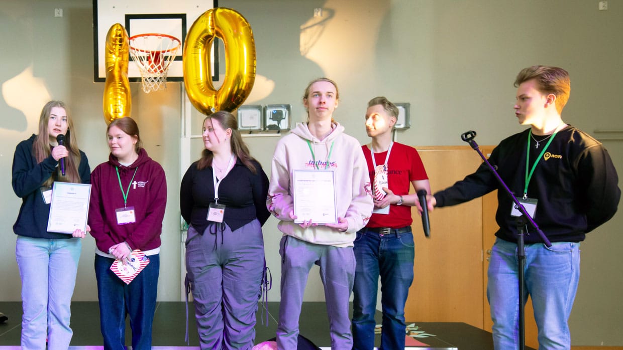 Neljä nuorta ja aikuinen miesoletettu seisovat rivissä. Hieman heidän edessään seisoo vielä yksi nuori miesoletettu mikrofonia pidellen. Rivissä seisovilla nuorilla on kahdella kädessään kunniakirja. Kaikilla on yllään rennot vaatteet. Nuoret katsovat hieman toisiaan ja ympärilleen.