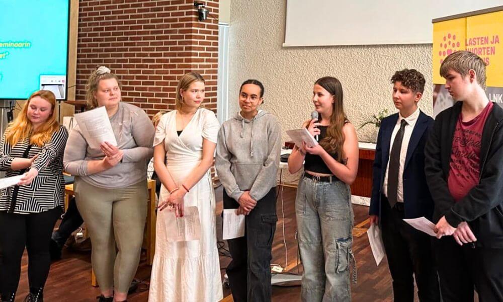 Nuoret seisovat rivissä. Yhdellä nuorella on kädessä mikrofoni.