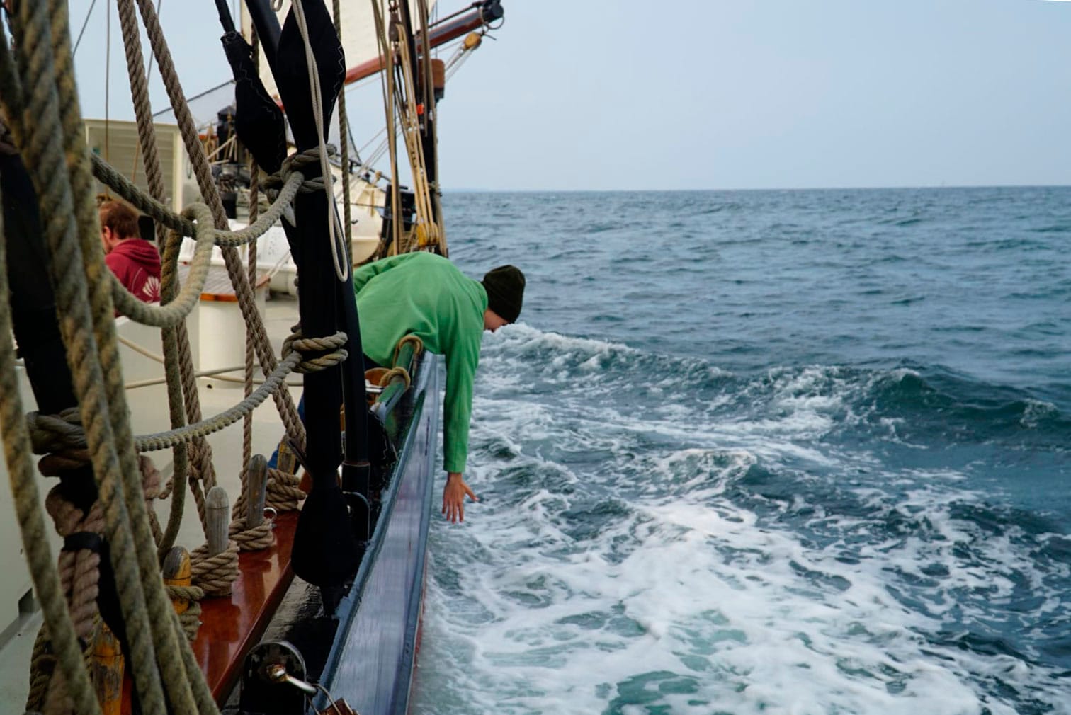 Henkilö kurottaa purjelaivan kannelta kohti aaltoja. Meri vaahtoaa alla, horisontissa ei näy muita aluksia tai maata. Hänellä on yllään vihreä college-paita ja päässä musta pipo.