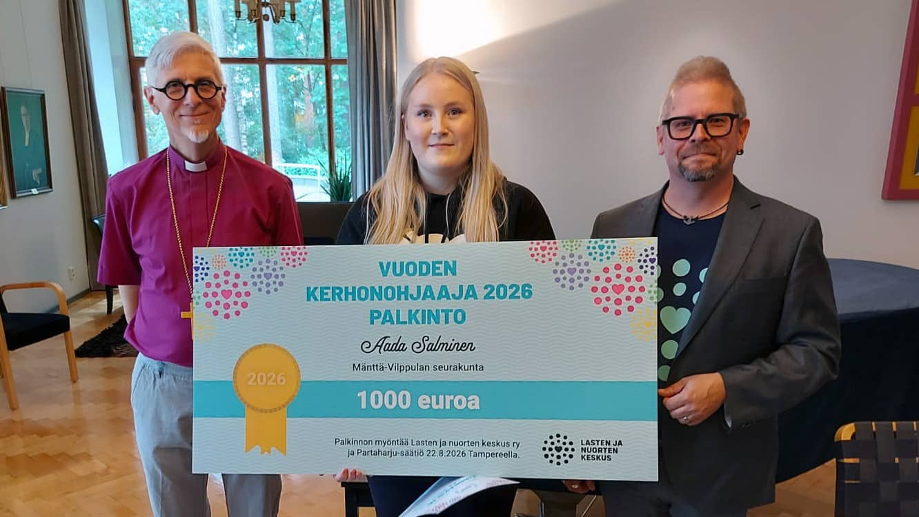 Kolme henkilöä seisovat. Keskimmäisellä henkilöllä on jättikokoinen shekki kädessä.